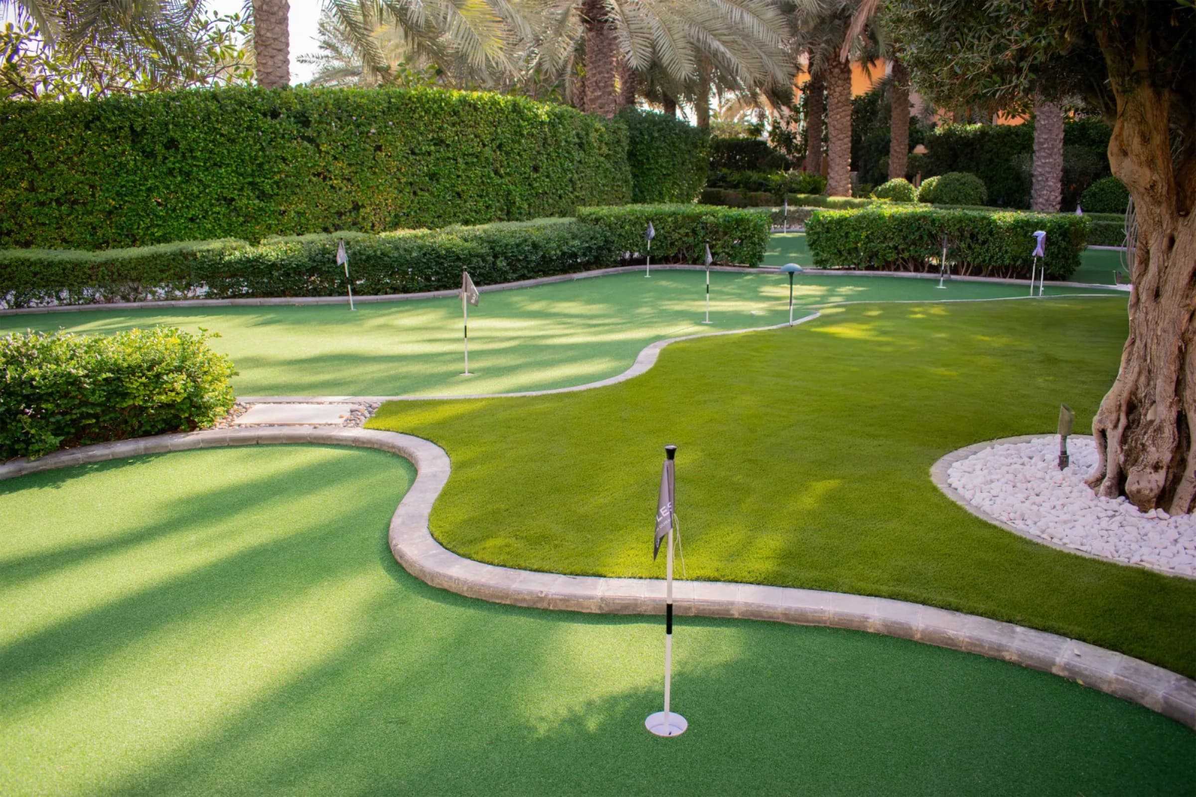 A contoured artificial putting green with several flagged holes, edged in stone and set among clipped hedges and palms in a Dubai garden.