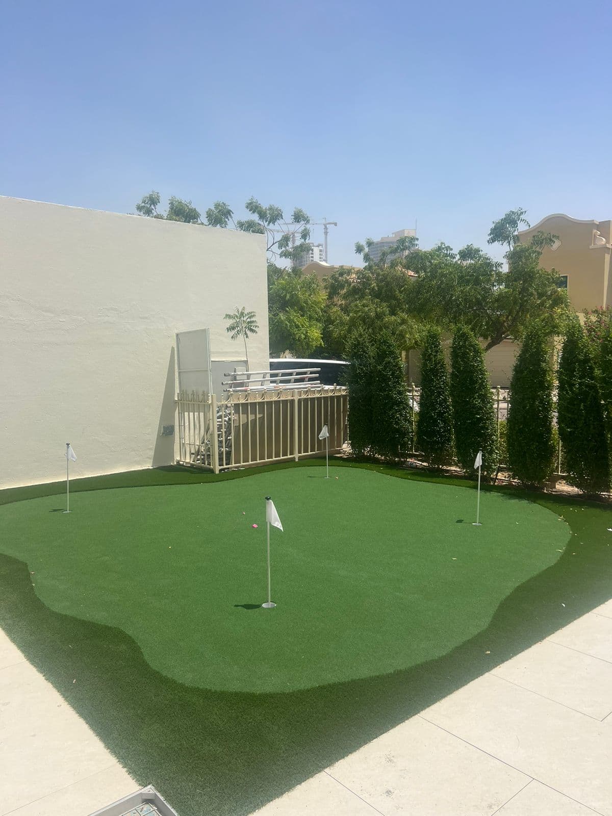 An artificial putting green with four flagged holes, edged by pale stone paving in a walled Dubai villa garden.