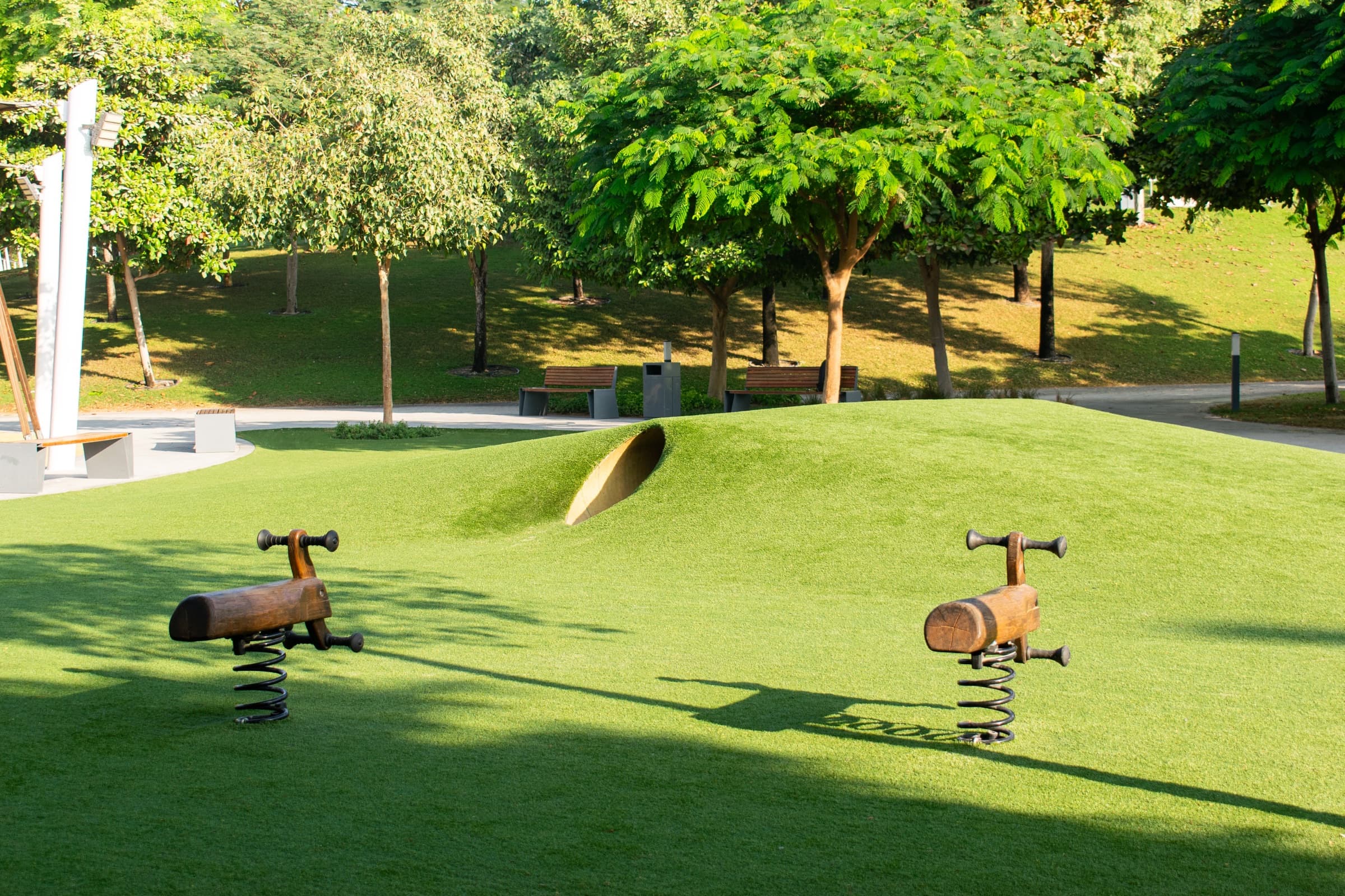 Two wooden spring rockers standing on a contoured artificial grass play area, with shaded trees and benches behind.